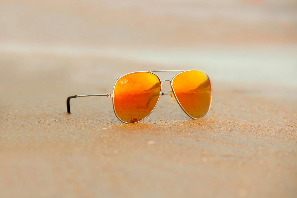 pexels-photo-249210-249210 Close-up of aviator sunglasses on sand with orange reflection, evoking a warm summer vibe.
