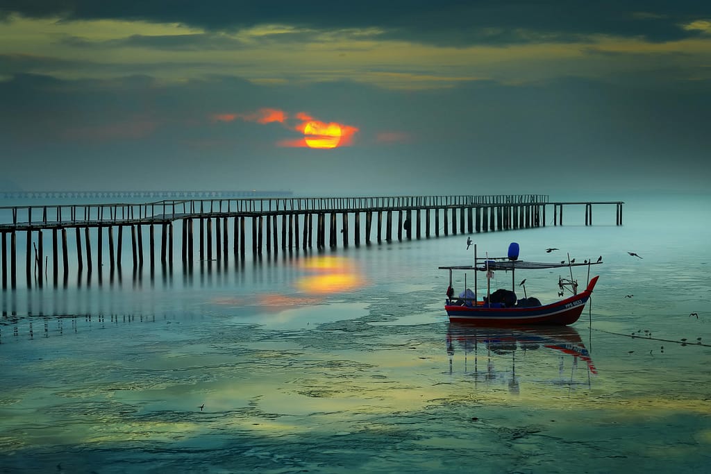 Serene sunrise view with boat and pier in George Town, Malaysia, reflecting vibrant colors on the sea.